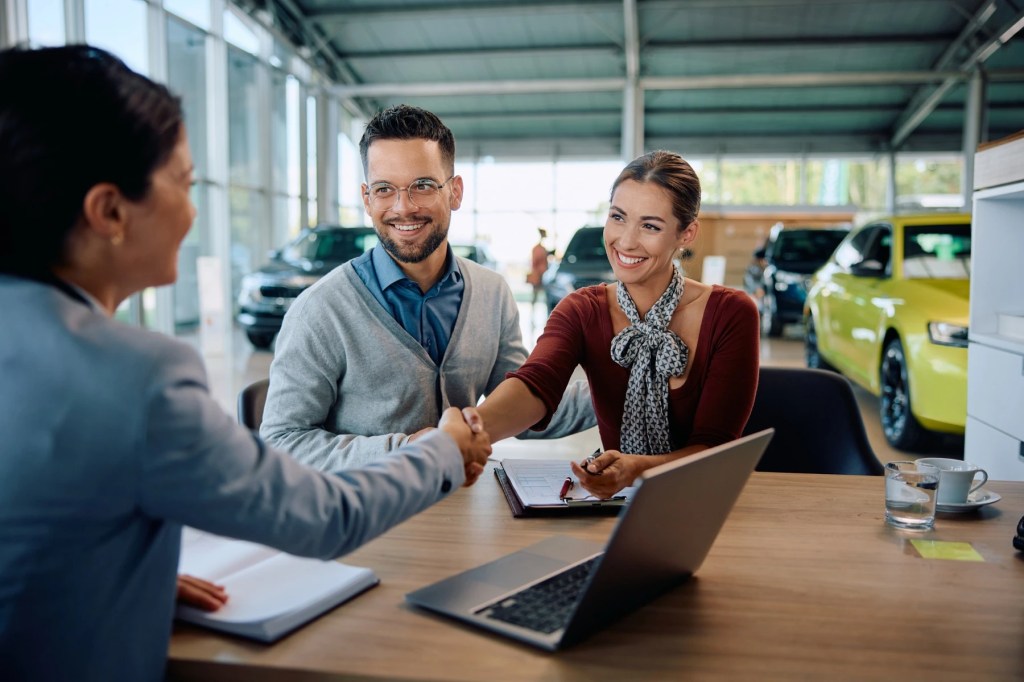 Couple shaking hands with a saleswoman at a car dealership desk