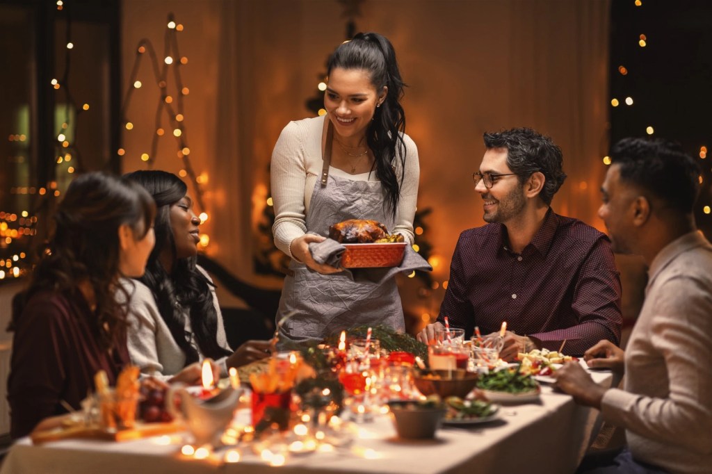 Woman bringing a roast to a candlelit holiday dinner table with friends