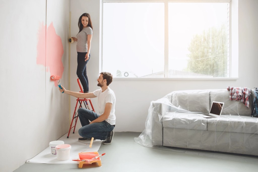 Couple painting an interior wall pink with furniture covered in plastic sheeting