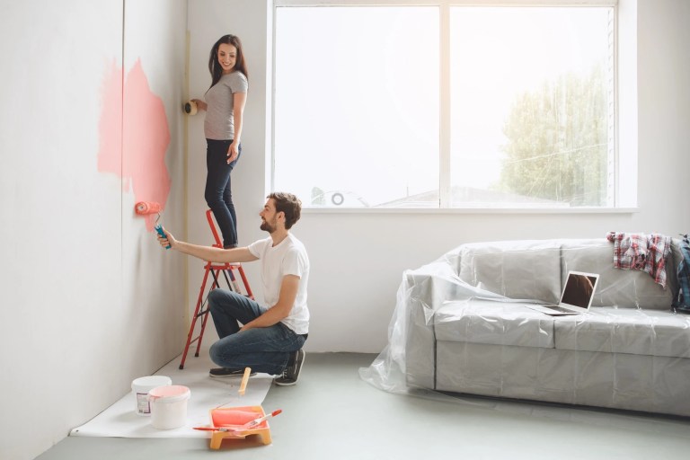 Couple painting an interior wall pink with furniture covered in plastic sheeting