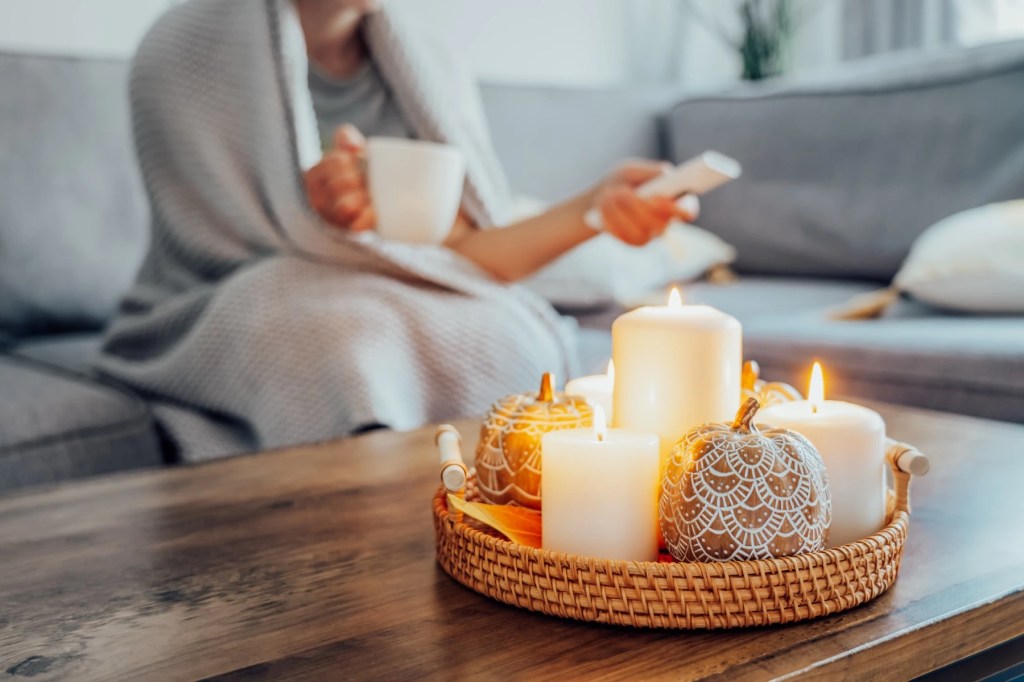 Lit candles and decorative pumpkins on a tray with a woman under a blanket behind